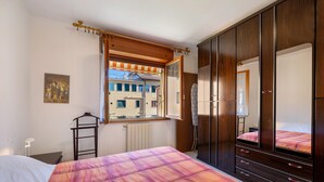 Ein Schlafzimmer mit Bett, Kleiderschrank, einem Fenster mit Vorhängen, einem Heizkörper und einem Bild an der Wand.
