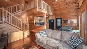 L’intérieur d’une cabane en bois comprend un escalier menant à une mezzanine, un salon avec un canapé et des fauteuils, et un coin cuisine.