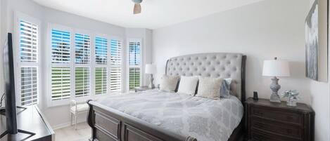 Une chambre à coucher avec un grand lit, deux tables de chevet, un ventilateur de plafond et une fenêtre avec des stores.