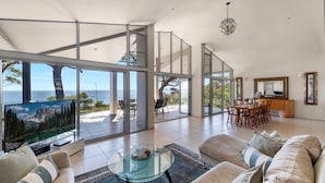 Un vaste espace de vie doté d’un grand mur de verre offrant une vue sur la mer, un coin repas avec des meubles en bois et une cuisine agrémentée d’un grand miroir.