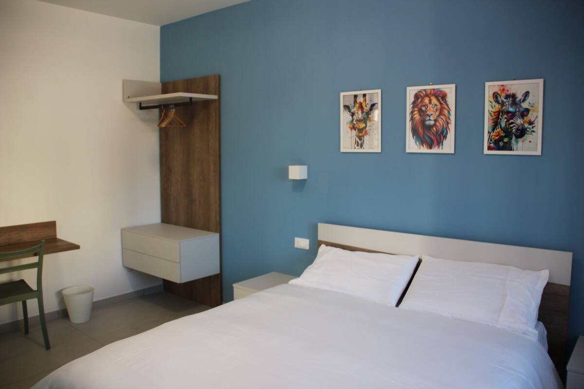 A hotel room with a bed, a desk, a chair, and artwork on the wall.