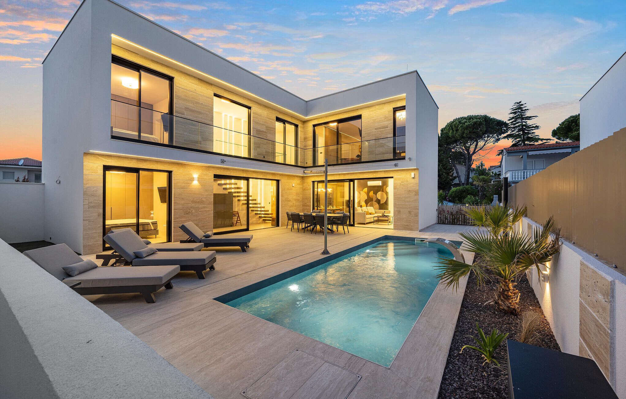 A modern two-story house with a swimming pool, outdoor seating, and a well-lit interior.