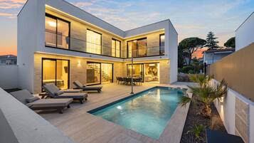 A modern two-story house with a swimming pool, outdoor seating, and a well-lit interior.