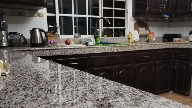 Una cocina con encimeras de granito, gabinetes de madera oscura, un ventanal con marcos blancos y varios electrodomésticos y utensilios sobre la encimera.