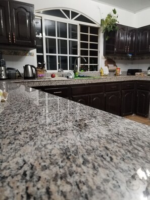 A kitchen with granite countertops, dark wooden cabinets, a window with white frames, and various kitchen appliances and items on the counter. - Boquete Guest House (Alto Boquete)