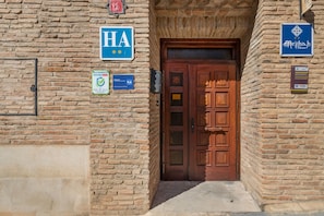 A brick building with a wooden door, signs indicating 'HA' and 'Alojamiento Seguro,' and a plaque with a rating of 9.6.