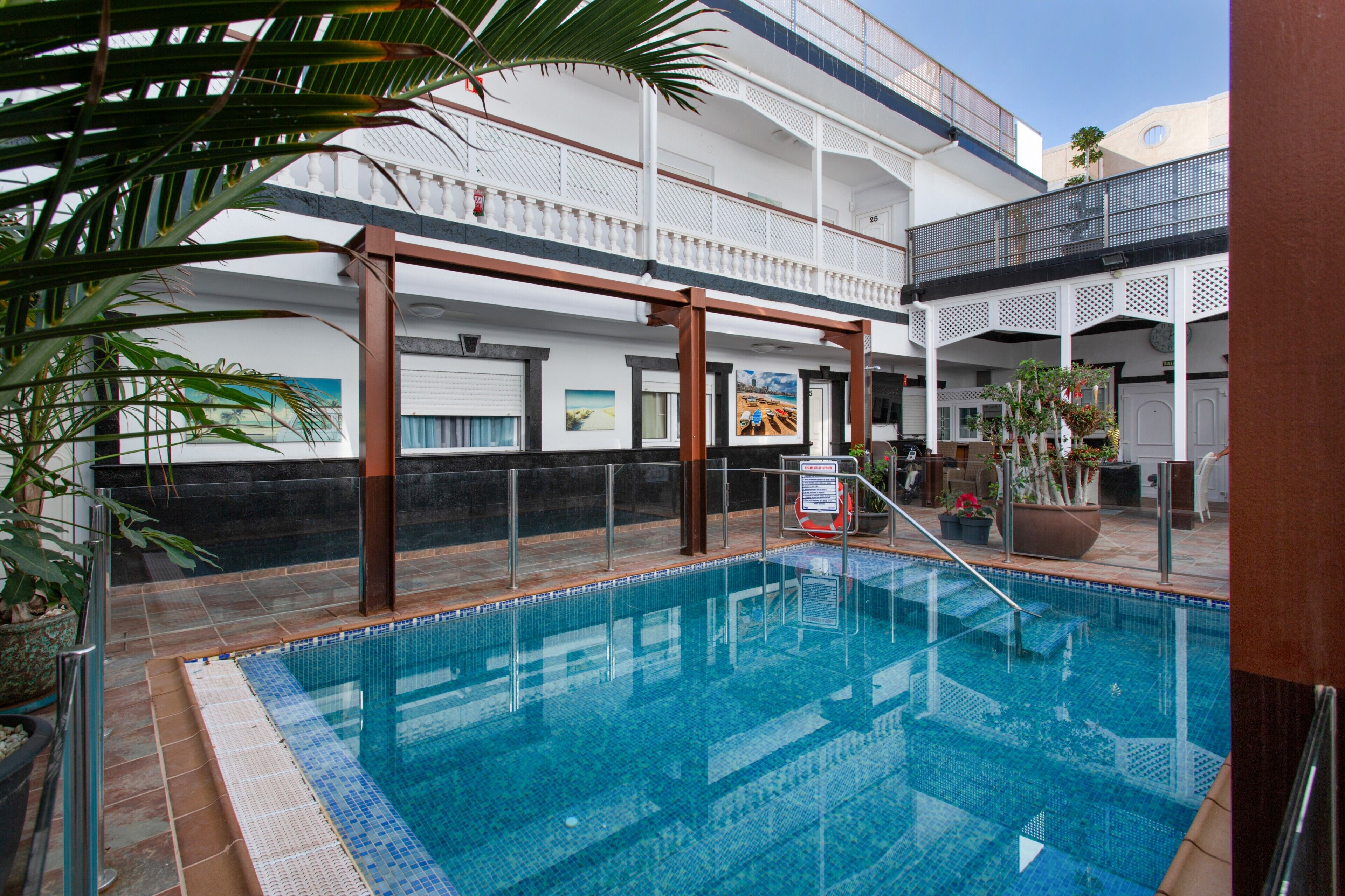 Une piscine intérieure avec un sol carrelé, entourée d’un bâtiment blanc doté de balcons et agrémentée de plantes en pot.