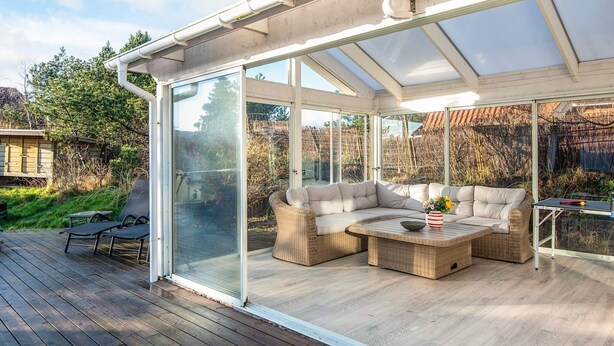 A covered outdoor seating area with a glass enclosure, wicker furniture, and a table with a vase of flowers. - Elegant Blavand Getaway- By Traum Ferienwohnungen (Blavand)