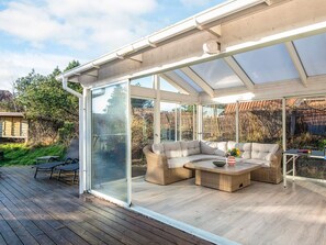 Un área de estar exterior techada con cerramiento de vidrio, muebles de mimbre y una mesa con un jarrón de flores.