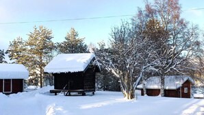 A wooden cabin with a snow-covered roof, surrounded by snow-covered trees and another cabin in the background. - 6 Person Holiday Home in Venjan (Venjan)