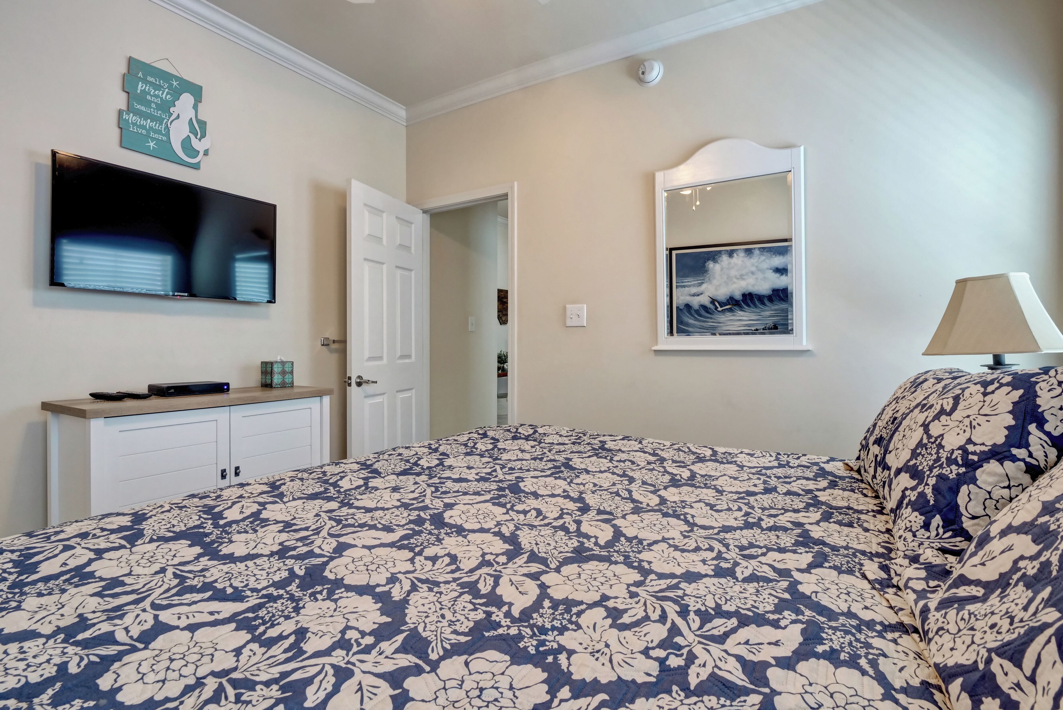 A bedroom with a floral-patterned bedspread, a mounted TV, a painting above the bed, and a door leading to another room.