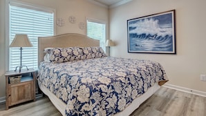 Une chambre avec un grand lit, deux tables de chevet, un ventilateur de plafond et un tableau représentant des vagues.