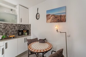 Una cocina con microondas, cafetera y lavabo; una mesa redonda con sillas; un reloj de pared; y una pintura con una escena de playa.