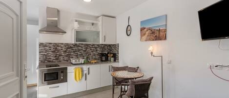 A compact kitchen with a microwave, toaster, and coffee maker, a dining area with wicker chairs, a ceiling fan, and a flat-screen TV mounted on the wall.
