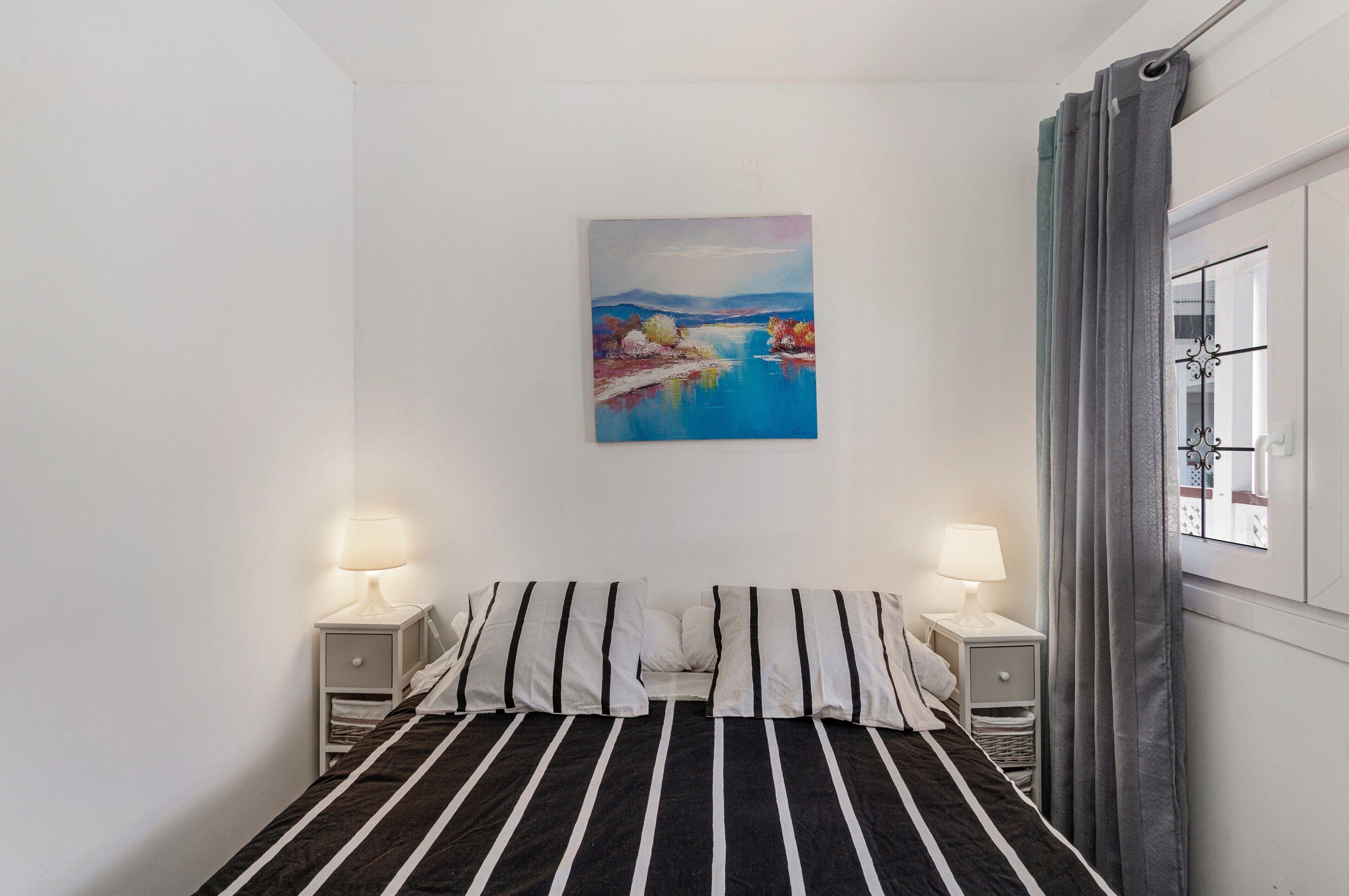 Een slaapkamer met een bed, twee nachtkastjes met lampen, een schilderij aan de muur en een raam met gordijnen.