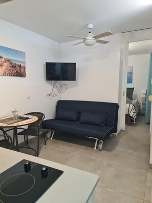 Een compacte leefruimte met een bank, een aan de muur bevestigde televisie, een eethoek met een tafel en stoelen, en een kitchenette met een kookplaat.