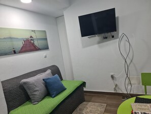 Une pièce avec un canapé gris, un plaid vert, un tableau représentant une jetée, un téléviseur fixé au mur et un fauteuil vert.
