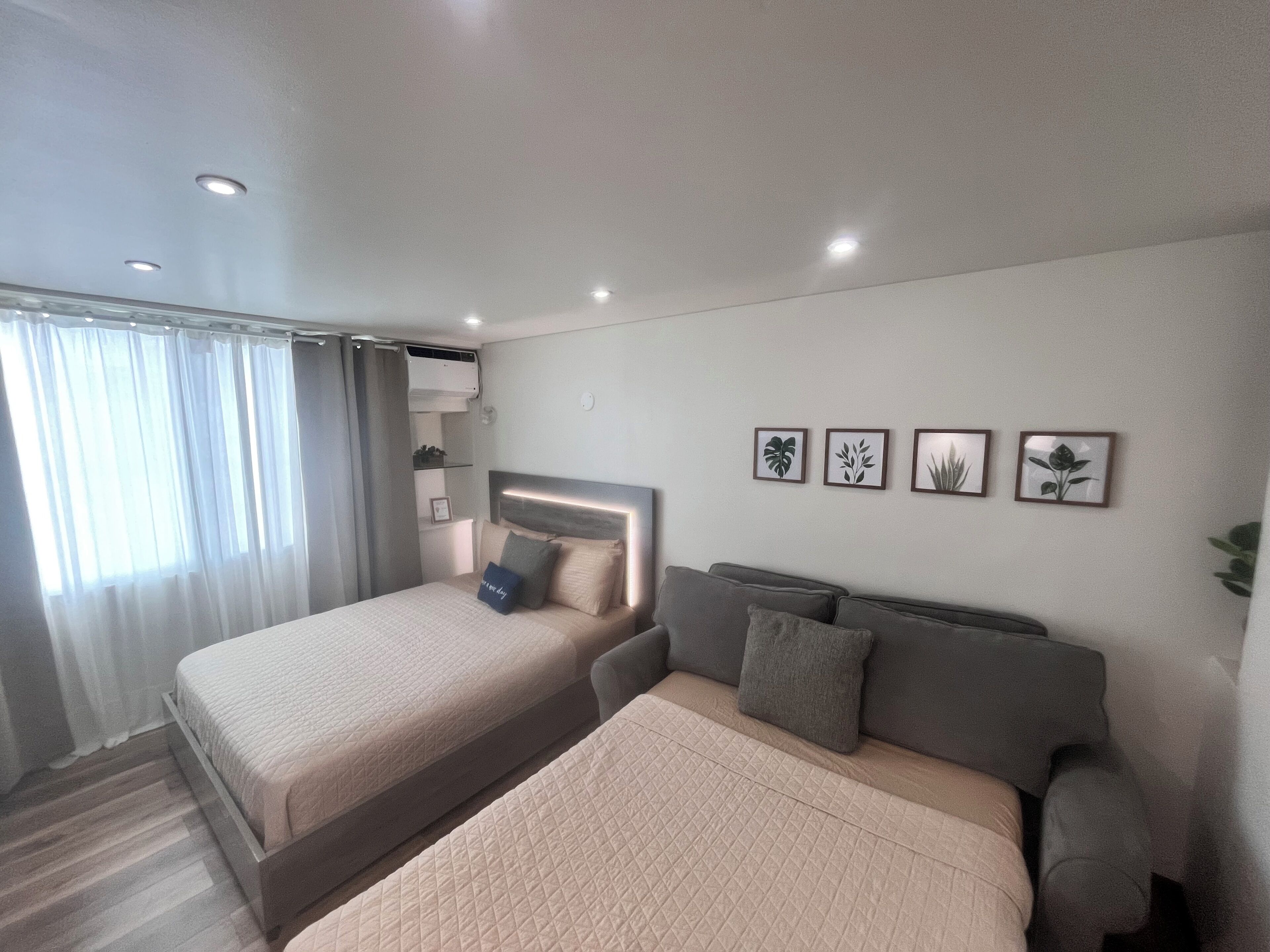 A modern bedroom with a large bed, a smaller sofa, and a wall-mounted TV. There are four framed plant pictures on the wall.