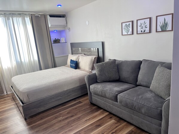 Ein modernes Schlafzimmer mit einem Bett, einem grauen Sofa, Holzböden und einem Deckenventilator.