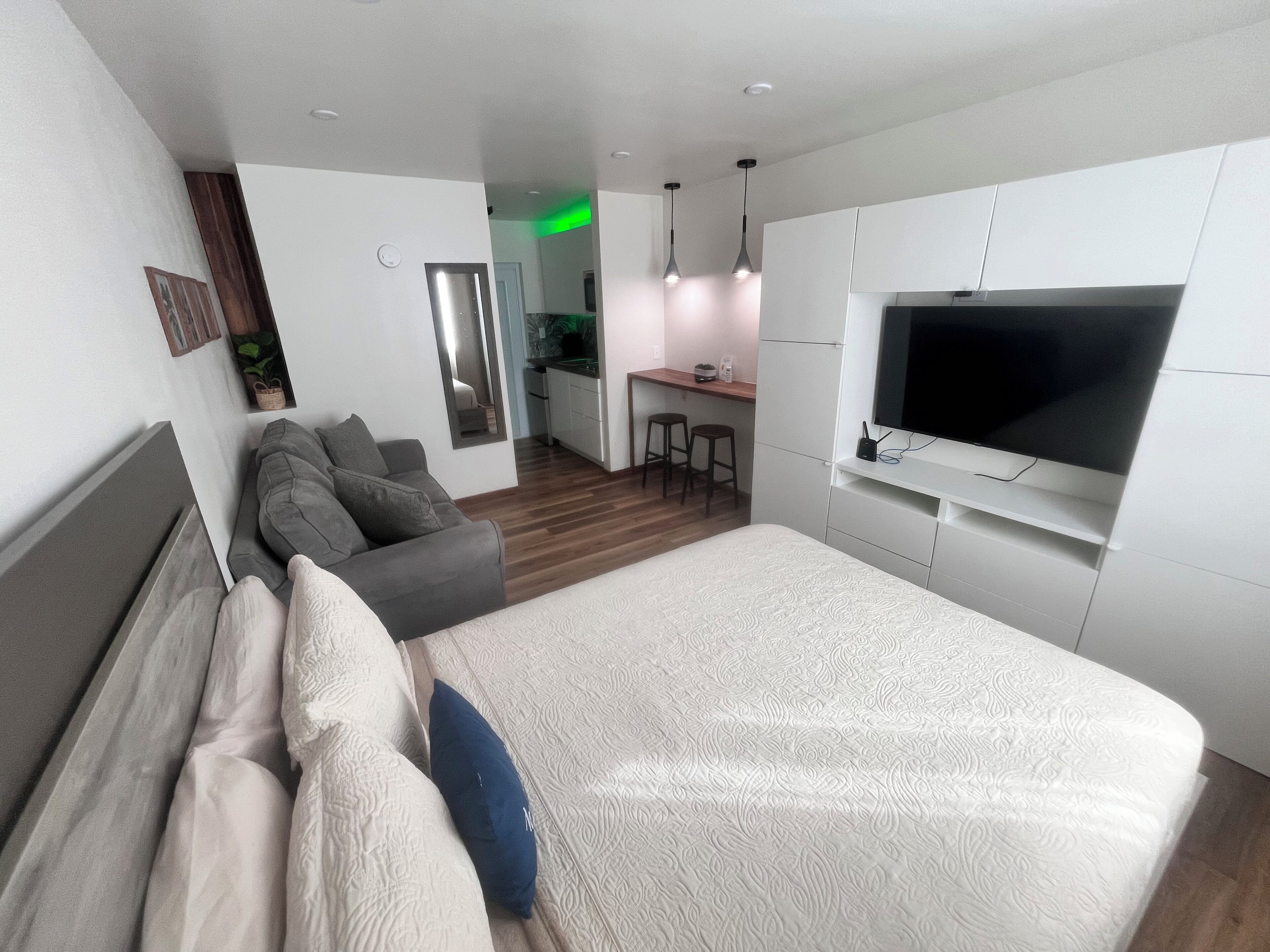 A modern bedroom with a large bed, a grey sofa, a TV mounted on the wall, and a small kitchenette in the background.