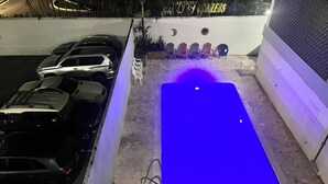 A swimming pool with a blue light, surrounded by parked cars and a white wall with decorative elements.