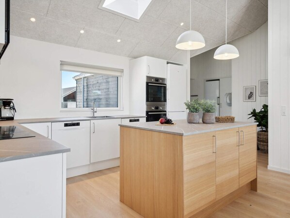 Une cuisine moderne avec des meubles blancs, un îlot central en bois, des luminaires suspendus et une vue sur l’extérieur.