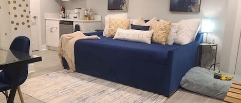 A modern bedroom with a large bed, a desk, and a kitchenette in the background.