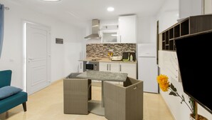 Cocina compacta con microondas, estufa y muebles altos blancos. Hay una mesa de comedor con sillas de mimbre y un televisor montado en la pared.