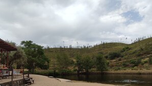 A sandy beach area with a small pavilion, a river, and a hilly landscape in the background.