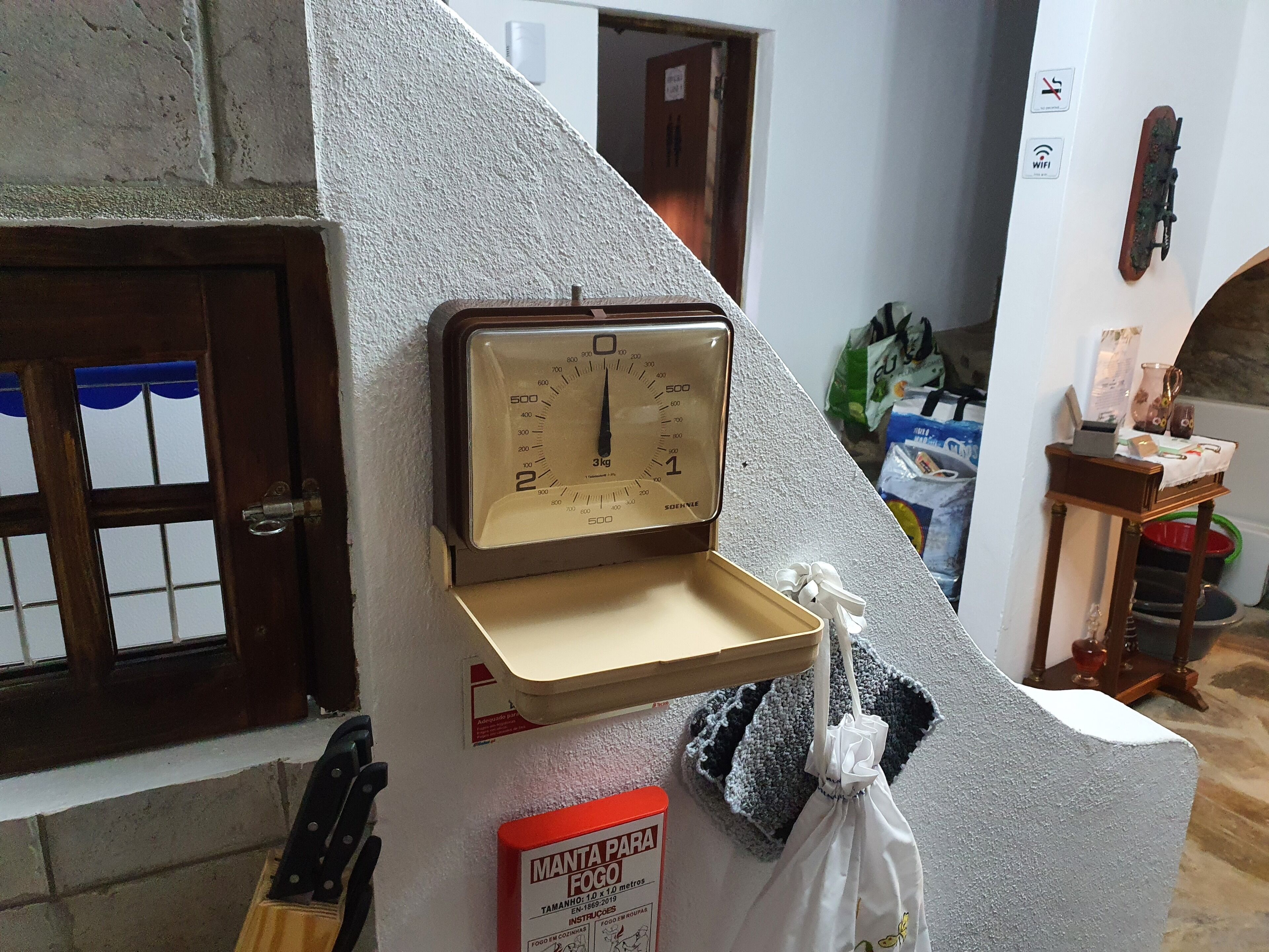 Una cocina con una balanza, una caja roja etiquetada “MANTA PARA FOGO” y una bolsa de tela colgando.