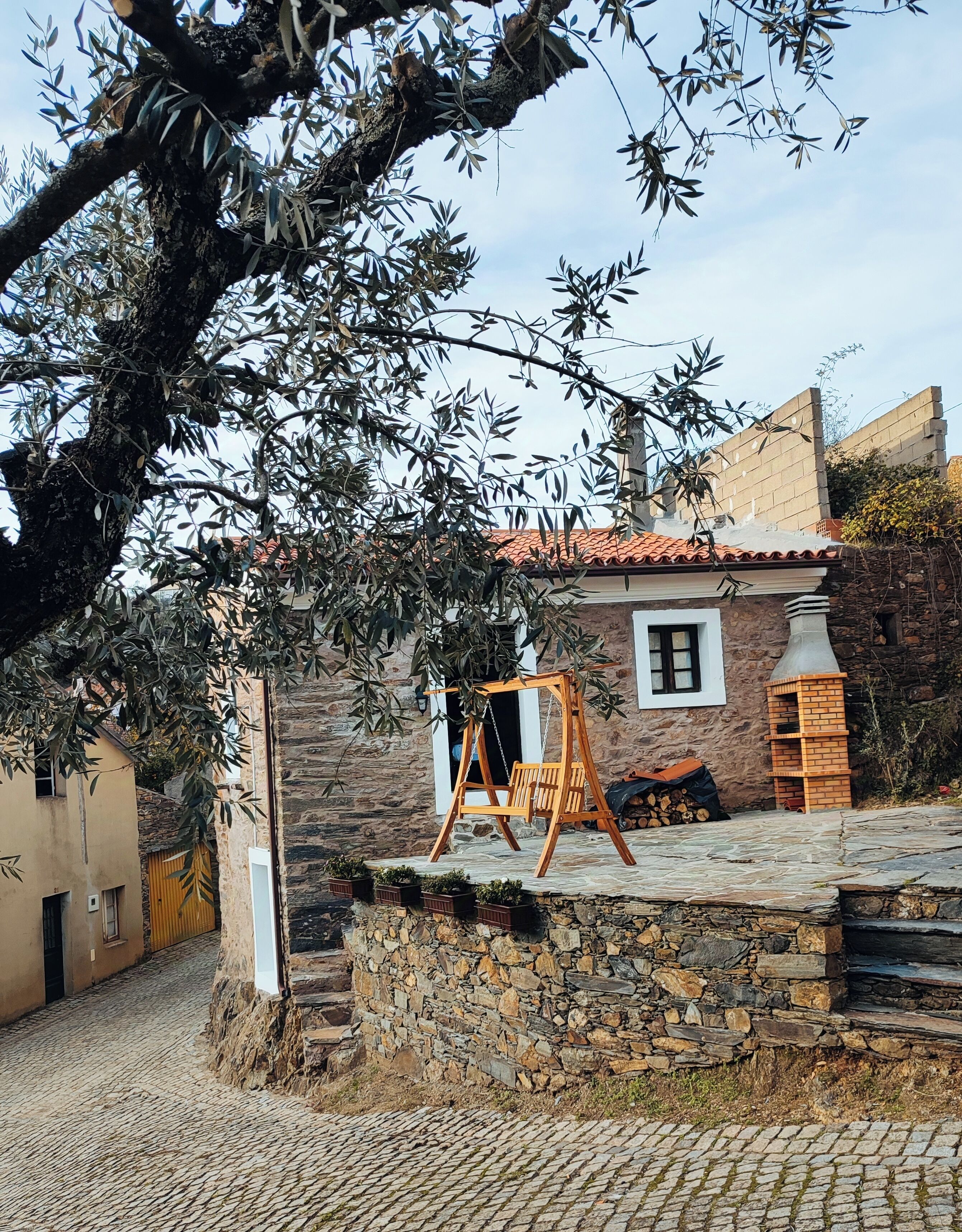A stone building with a wooden swing set in front, a stone wall, and a tiled roof.