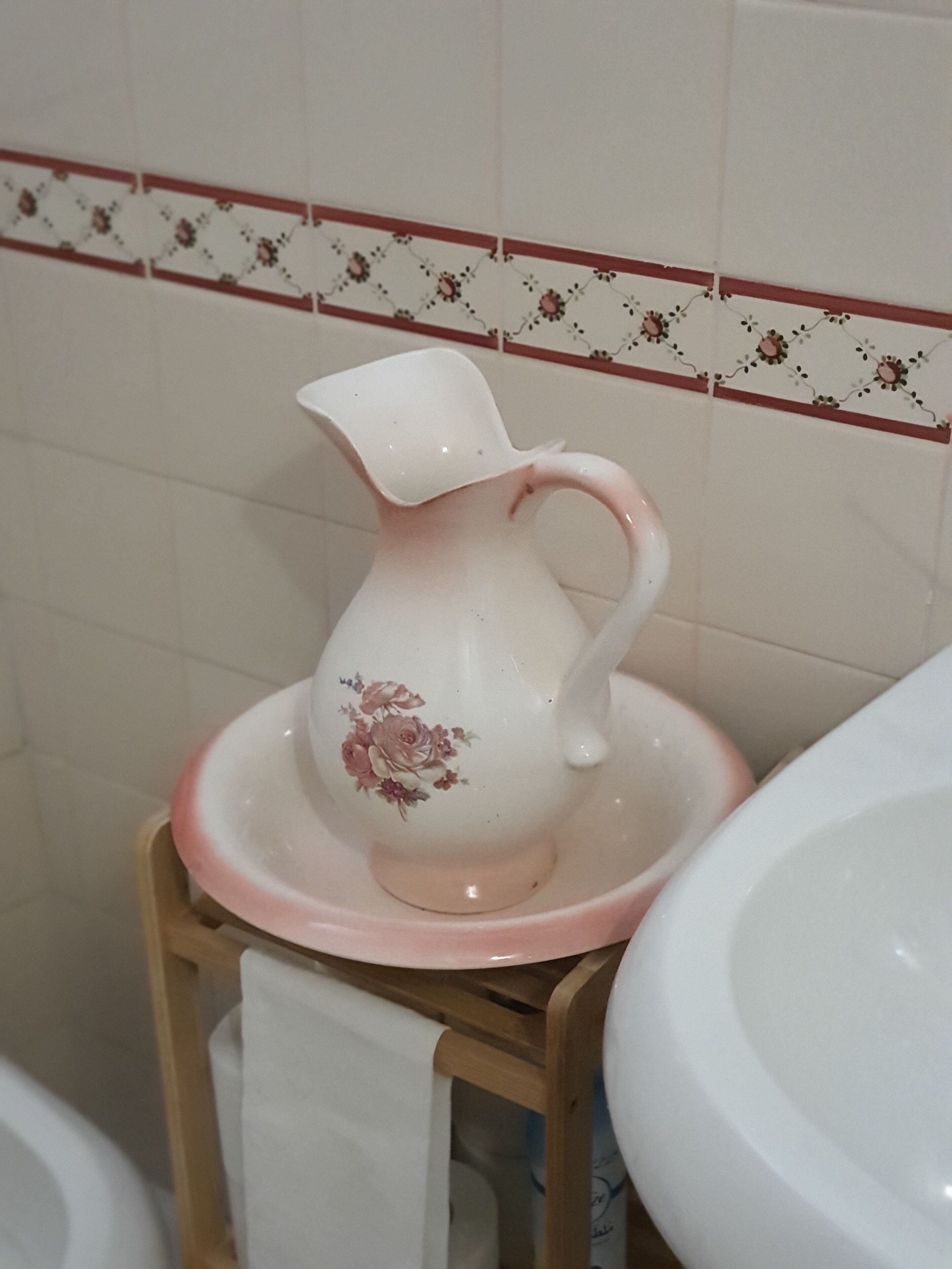 A bathroom with a white ceramic pitcher on a wooden stool, a white sink, and a tiled wall with a floral border.