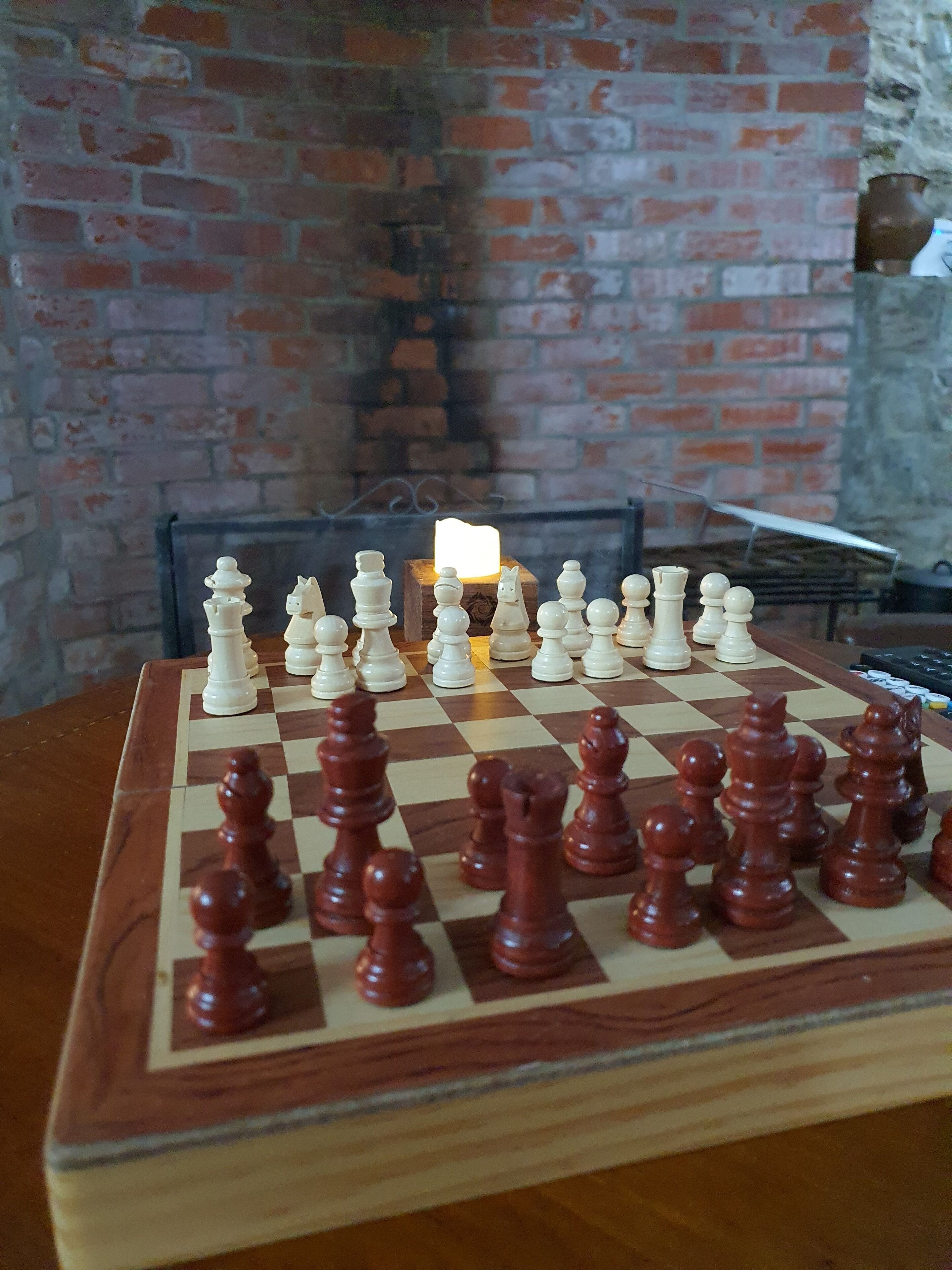 Un échiquier avec les pièces disposées pour une partie, posé sur une table en bois dans une pièce dont un mur est en briques et où une bougie allumée se trouve à l’arrière-plan.