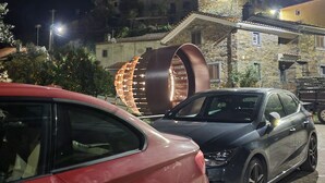 A parking area with two cars, a stone building with a sloped roof, and a large illuminated structure in the background.