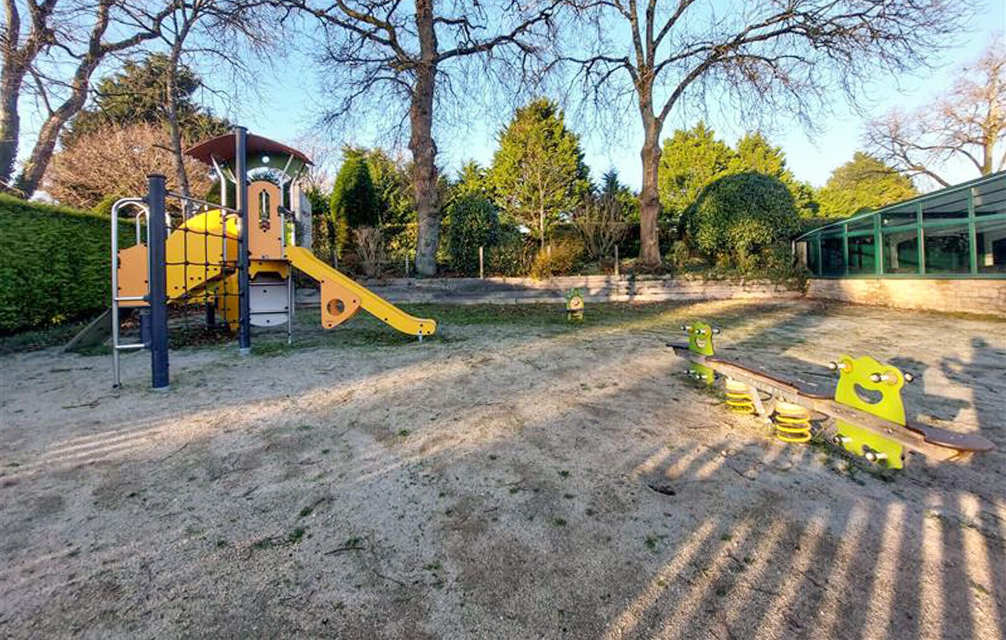 A playground with a slide and climbing structures, surrounded by trees and a building in the background.