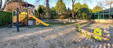 A playground with a slide and climbing structures, surrounded by trees and a building in the background.