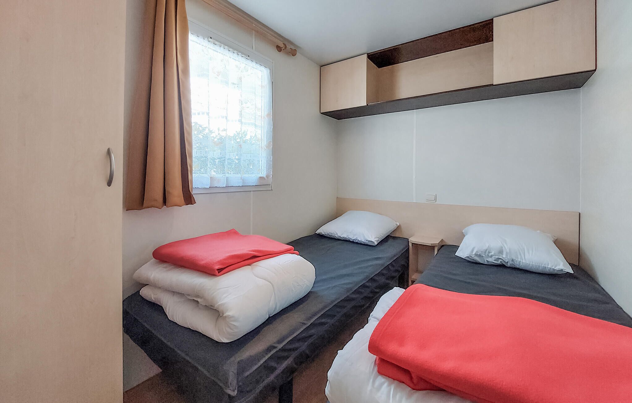 A small, compact room with two single beds, a window with curtains, and a built-in shelf above the beds.