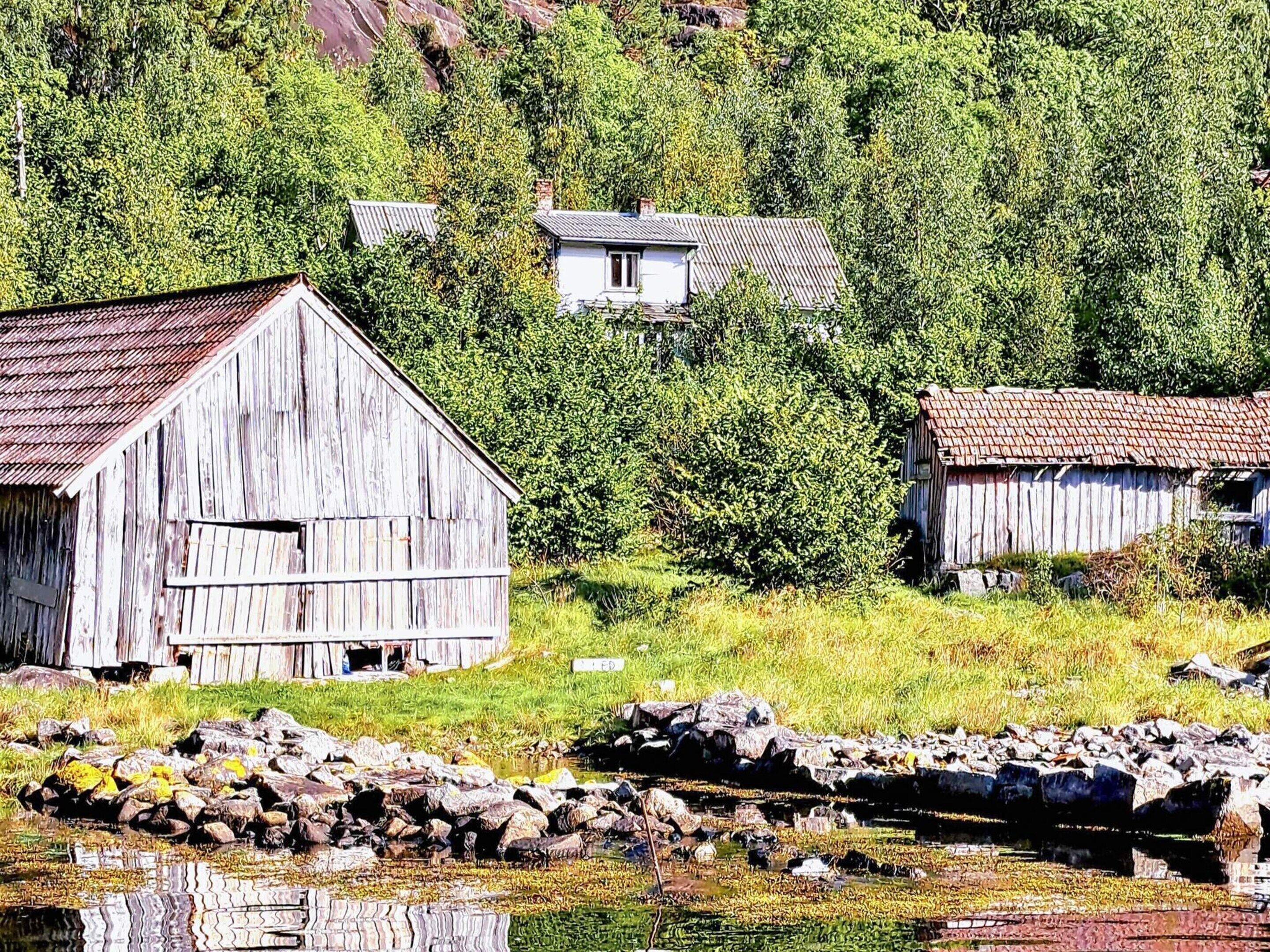 A rustic wooden barn with a metal roof, surrounded by lush greenery and a rocky shoreline.