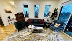 Ein Wohnzimmer mit einer schwarzen Ledersofa, einem grünen Sessel, einem weißen Couchtisch und einem großen, flauschigen Teppich. Zwei Fenster mit Vorhängen, eine Pflanze und eine Holzkommode sind ebenfalls vorhanden.