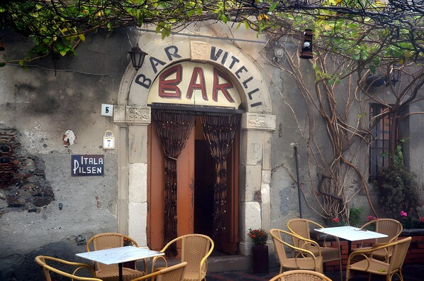 Un área de bar al aire libre con sillas y mesas de mimbre, un letrero que dice 'BAR VITELLI' y un cartel de 'PITALA PILSEN' en la pared.