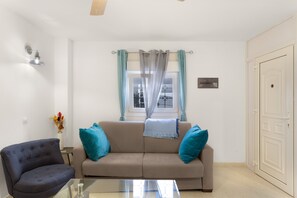 A living room with a sofa, a chair, a glass table, a window with curtains, a wall-mounted light, and a door.