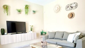 Ein Wohnzimmer mit einem Flachbildfernseher, einem grauen Sofa, einem Korbtisch und mehreren Kübelpflanzen auf einer weißen Ablage.