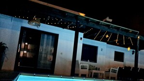 Ein Poolbereich mit freiem Blick auf den Pool, eine überdachte Sitzgelegenheit mit Stühlen und ein Gebäude mit großen Fenstern und einer Tür.
