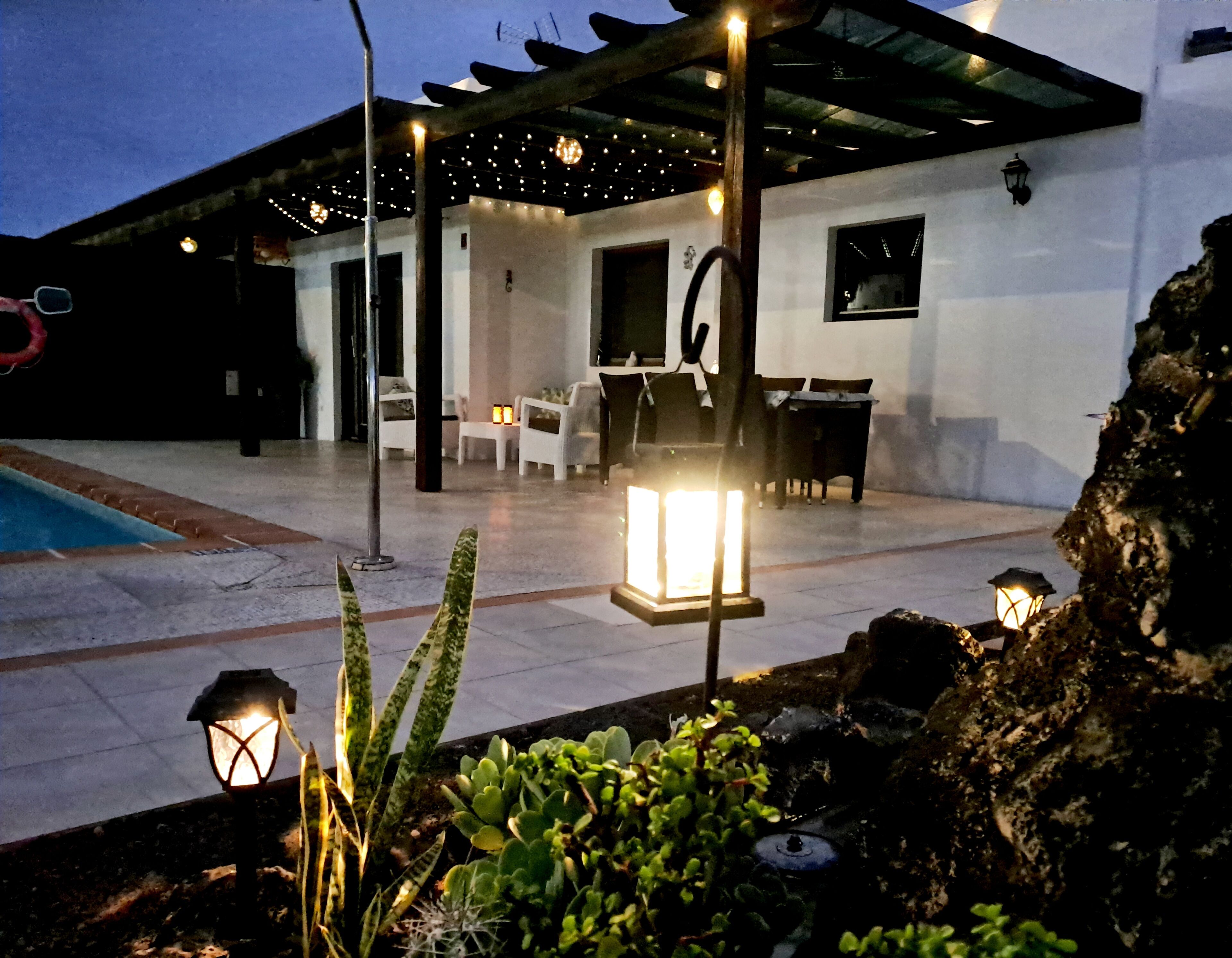A covered outdoor patio with seating, a pool, and a well-lit garden area.