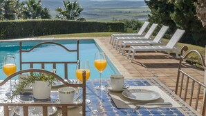 A poolside dining area with a table set for two, two glasses of orange juice, a potted plant, and a view of the mountains. - Villa 'Lirio' with Mountain Views, Wi-Fi, and Air Conditioning (Benalup-Casas Viejas)