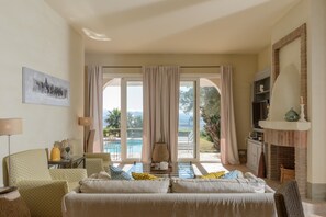 Una sala de estar con sofá, televisión y chimenea. Se aprecia una ventana grande con cortinas y vistas a una piscina y vegetación.