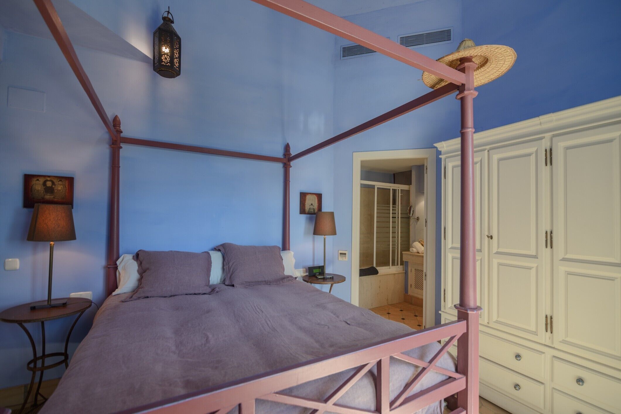 Un dormitorio con cama con dosel, una mesita de noche, una lámpara, un armario y una puerta que da a otra habitación.