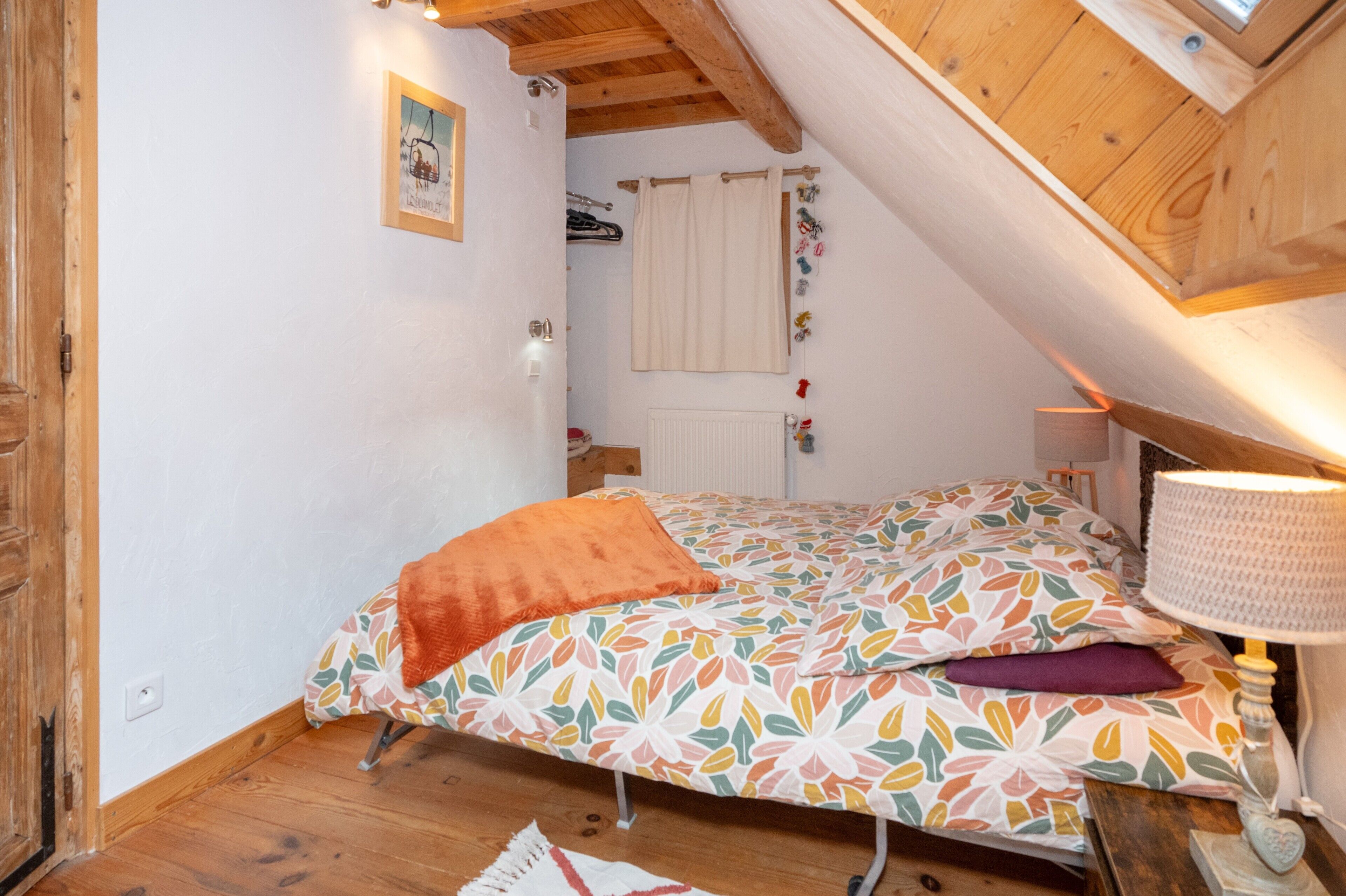A bedroom with a sloped ceiling, a bed with a colorful floral bedspread, a wooden floor, and a wall-mounted radiator.