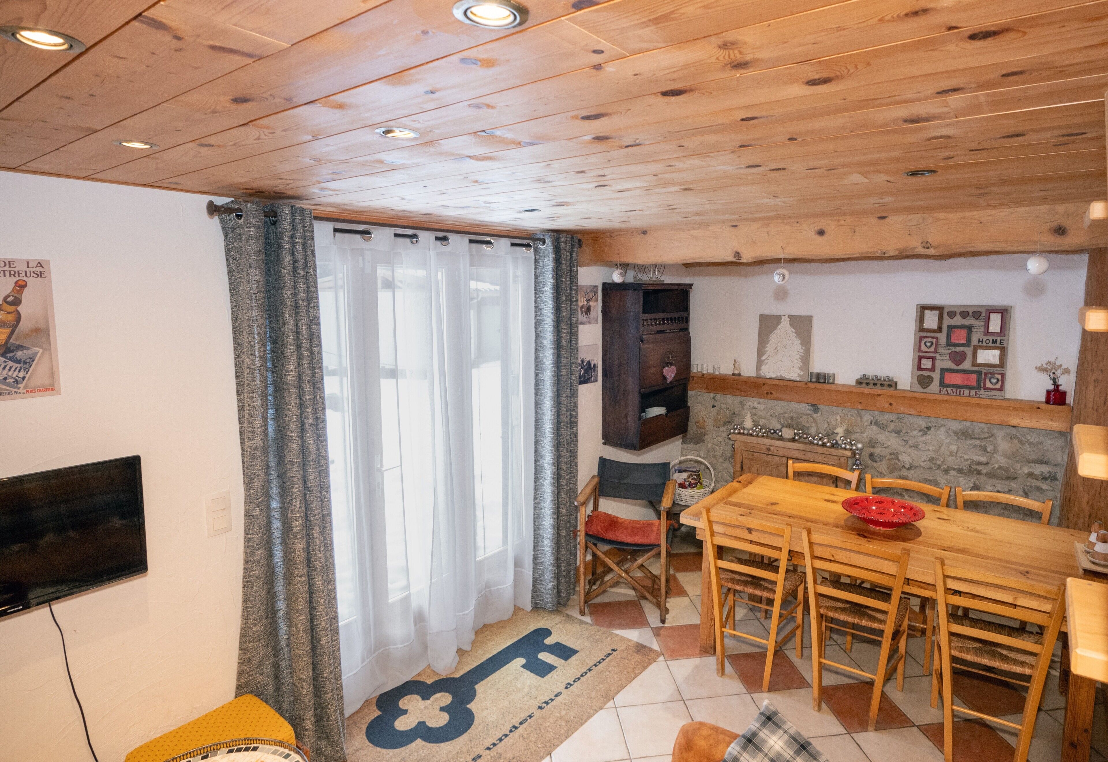 Ein gemütliches Wohnzimmer mit Holzbalkendecke, einem an der Wand befestigten Fernseher, einer Essecke mit Tisch und Stühlen und einem großen Fenster mit Vorhängen.