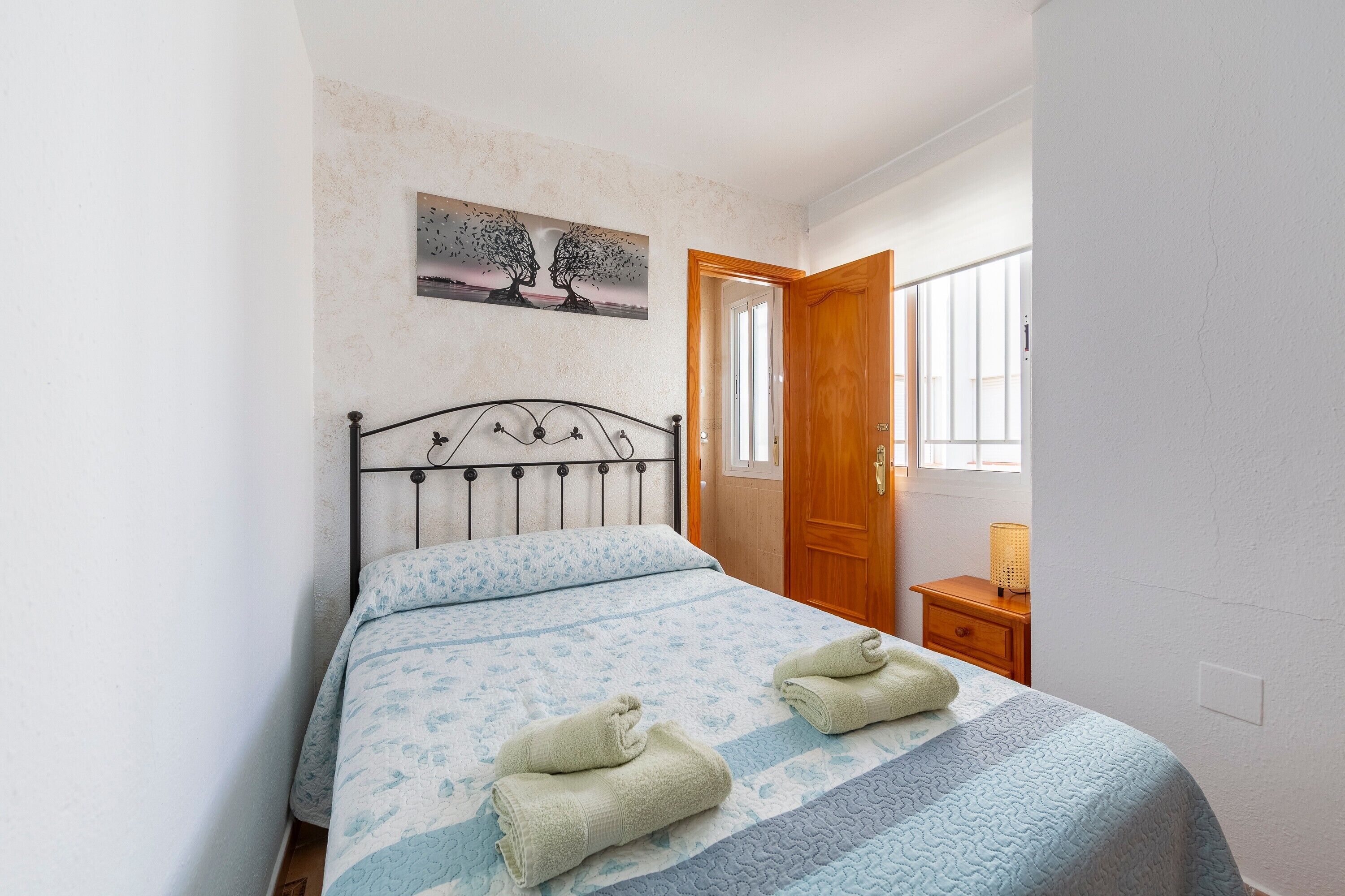 A bedroom with a bed, two towels, a nightstand, a lamp, a door, and a painting on the wall.